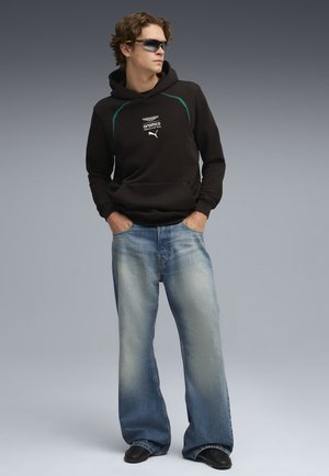 Ung mand iført sort Aramco Formula One hættetrøje, blå bukser med vide ben, sorte sko og mørke solbriller, stående med hænderne i lommerne.