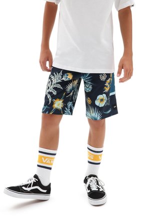 Dziecko ubrane w białą koszulkę, czarne krótkie spodenki w kwiatowy wzór, białe skarpetki Vans z żółtym paskiem i czarne trampki Vans, stojące na białym tle.