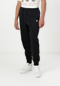 Schwarze Jogginghose mit elastischem Bund, verstellbarem Kordelzug, seitlichen Taschen und weißem Nike-Logo; kombiniert mit weißen Sneakers.