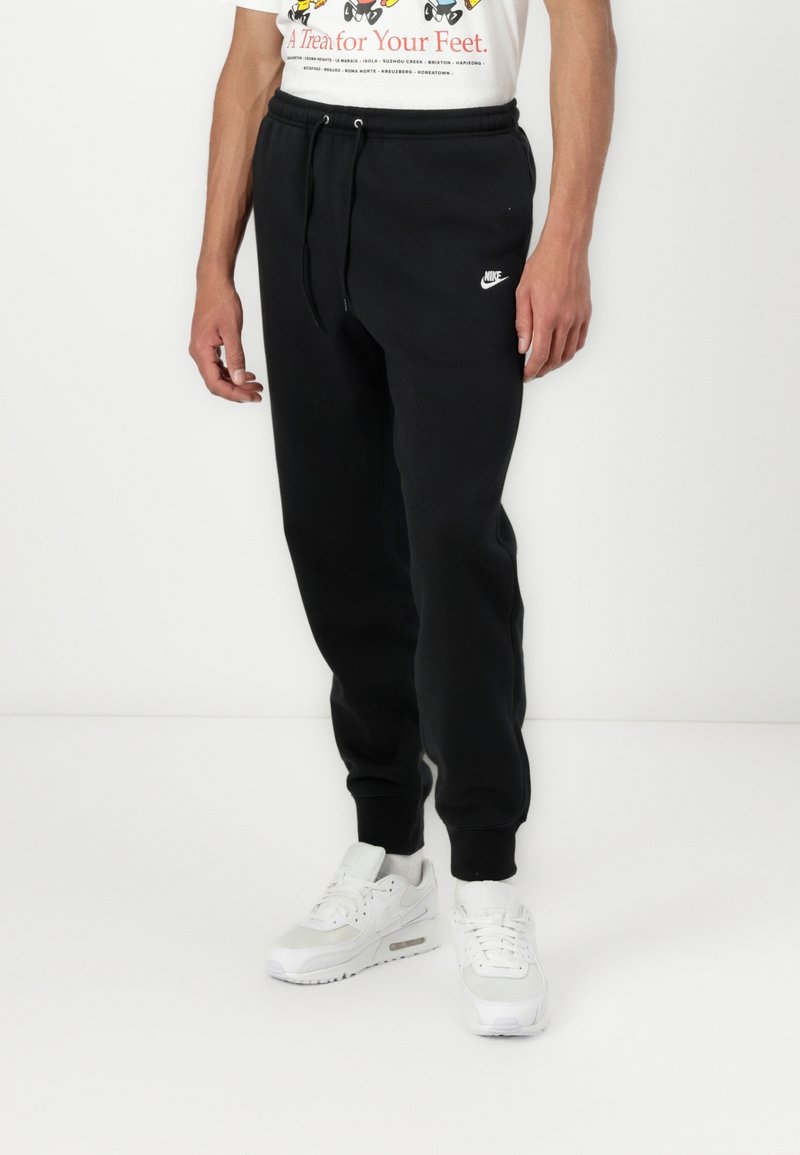 Schwarze Jogginghose mit elastischem Bund, verstellbarem Kordelzug, seitlichen Taschen und weißem Nike-Logo; kombiniert mit weißen Sneakers.