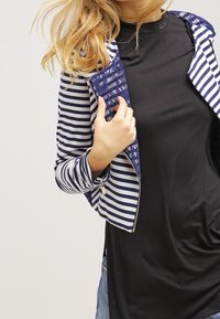 Modrý a bílý pruhovaný jacket s přiléhavým designem, s zipovým uzávěrem a vzorovými akcenty. Nosič je oblečen přes černou košili.