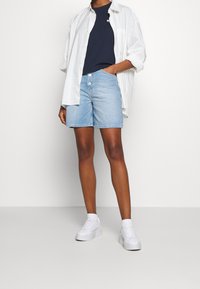 Ljust blå denimshorts med hög midja, två knappar, i kombination med en marinblå t-shirt och en lös vit skjorta med knappar. Vita sneakers.