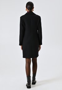 Cappotto di lana nera con vestibilità su misura, colletto a rever e maniche lunghe. Presenta un taglio al ginocchio e un design minimalista.