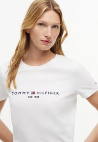 Femme aux longs cheveux blonds portant un t-shirt blanc Tommy Hilfiger avec le logo et le texte "Est. 1985" sur la poitrine, face à l'avant.