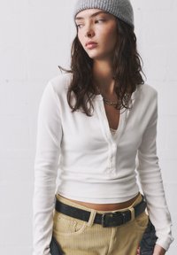 Valkoinen ribattu pitkähihainen paita napitusläpällä, yhdistettynä vaaleanbeigeihin corduroy-housuihin ja mustaan vyöhön. Harmaa neulottu pipo päässä.