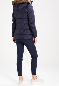 Donna che indossa una giacca trapuntata blu navy con cappuccio foderato in pelliccia, pantaloni slim blu navy e sneakers color beige chiaro, in piedi contro uno sfondo bianco.