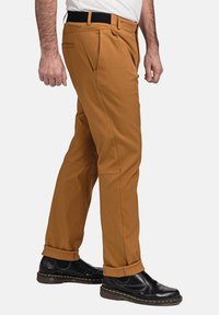 Braune Hosen mit glatter Textur, im entspannten Schnitt, mit Falten und einem schwarzen Bund, kombiniert mit schwarzen Schuhen.