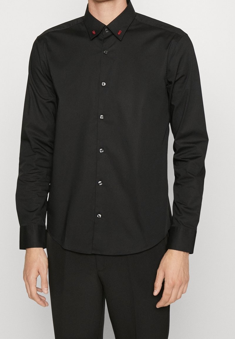 Homme portant une chemise noire à manches longues avec des détails brodés rouges sur le col, assortie à un pantalon noir.