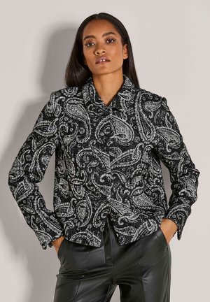 Vrouw met lang donker haar, gekleed in een zwart-wit paisley-gepatroneerde button-up jack en zwarte leren broek, met de handen in de zakken.