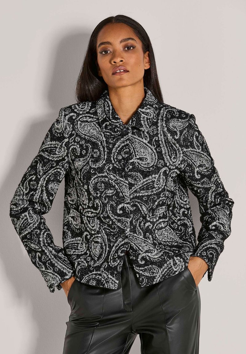 Frau mit langen dunklen Haaren, die eine schwarz-weiße Jacke mit Paisleymuster und schwarze Lederhosen trägt, mit den Händen in den Taschen.