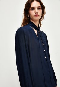 Blusa blu navy con design plissettato, caratterizzata da un collo con laccetto e dettagli con bottoni dorati. Realizzata in tessuto leggero e morbido.