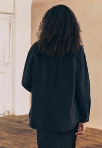 Mustat farkkukangas ylisuurissa paidassa on kaareva helma, nappihihansuut ja takana sauma. Pehmeä pinta, jossa on hieman haalistunut viimeistely.