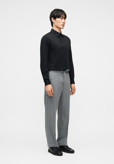 Νέος άνδρας στέκεται φορώντας μαύρο πόλο με μακριά μανίκια, γκρι παντελόνι με ρίγες και κορδόνια, καθώς και μαύρα παπούτσια, σε απλό λευκό φόντο.