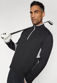 Mustas zip-up golfipullover hallide aktsentide ja tekstuuriga kangaga, koos valgete golfikinnastega. Golfikepp puusal.