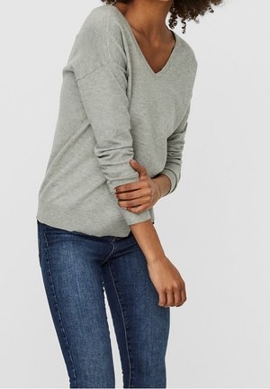 Femme portant un pull gris clair à col en V et un jean bleu, debout avec un bras croisé tenant le coude opposé.