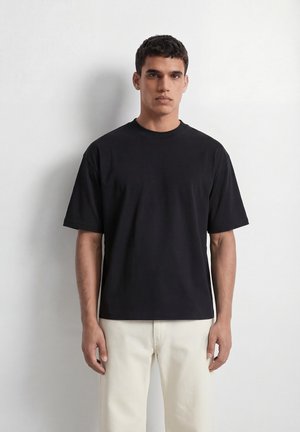 Mann steht an einer weißen Wand, trägt ein schlichtes schwarzes T-Shirt mit kurzen Ärmeln und helle Hosen, blickt geradeaus mit neutralem Gesichtsausdruck.