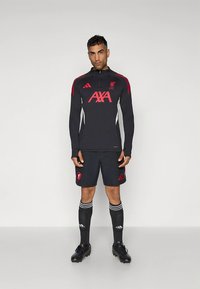 Juodas sportinis kostiumas su raudonomis detalėmis, turi užtrauktuku užsegamą viršutinę dalį, logotipą ir šortus. Komplekte yra dryžuotos kojinės ir juodi futbolo bateliai.