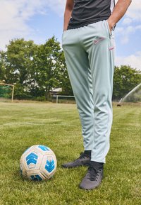 Lyseblå Nike træningsbukser med et pink logo, båret med sorte støvler, står ved siden af en mønstrede fodbold på græs.