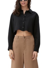 Vrouw in een zwarte cropped blouse met knopen, beige high-waisted broek en zwarte zonnebril, staande tegen een witte achtergrond.