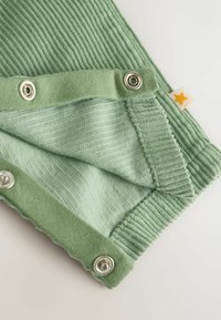 Tissu vert clair avec une texture à côtes, doté de boutons-pression. Comprend une petite étiquette avec une étoile jaune sur le bord.