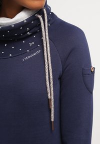 Felpa con cappuccio blu navy e colletto alto, con motivi geometrici bianchi e foderatura grigio chiaro. Include lacci intrecciati e una tasca laterale.