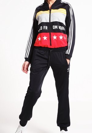 Henkilö, joka pukeutunut mustaan verkkatakkiin, jossa keltaisia, harmaita ja punaisia väripintoja, valkoiset raidat hihoissa, tähtiä edessä, ja valkoiset tennarit.