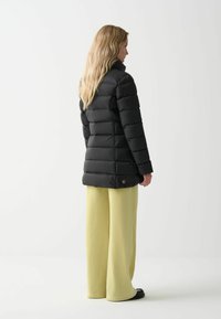 Zwarte pufferjack met hoge kraag en horizontale stiksels, gecombineerd met lichtgele wijde broek en zwarte schoenen. Minimalistisch ontwerp.