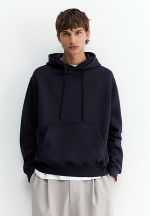 Giovane con capelli castani corti che indossa una felpa nera con cappuccio e pantaloni beige chiaro, in piedi davanti a uno sfondo bianco semplice.