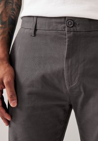 Męskie ciemnoszare spodnie wykonane z tkaniny mieszanki bawełny, wyposażone w zapięcie na guzik, boczne kieszenie oraz gładką fakturę. Widoczne szczegółowe przeszycia.