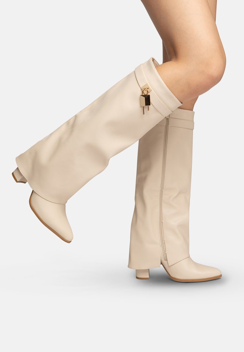 Beige leren laarzen tot aan de knie met lage blokhakken en een gouden hangslotdetail aan de zijkant, gedragen op gekruiste benen tegen een witte achtergrond.