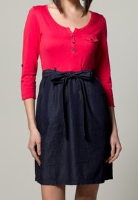 Czerwony top z długim rękawem i dekoltem henley oraz ciemnoniebieska spódnica dżinsowa z wiązaniem w talii i bocznymi kieszeniami. Gładka tekstura, casualowy design.