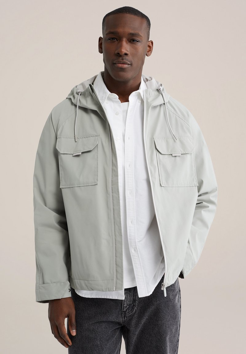 Lichtgrijze jas met een capuchon, voorzien van twee borstzakken, ritssluiting en elastische manchetten, gedragen over een wit overhemd met knopen.