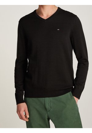 Homme portant un pull noir à col en V et un pantalon vert, une main dans la poche, sur un fond uni.