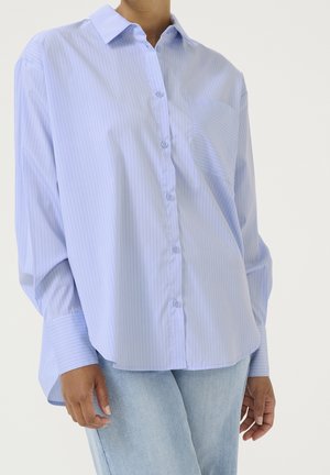 Personne portant une chemise ample bleu clair à boutons avec de fines rayures verticales et un jean bleu clair sur un fond uni.