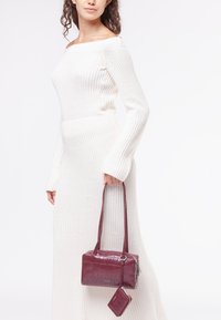 Borsa con stampa coccodrillo color bordeaux, caratterizzata da una texture liscia, una lunga tracolla e una piccola pochette attaccata. Indossata a mano contro un vestito bianco.