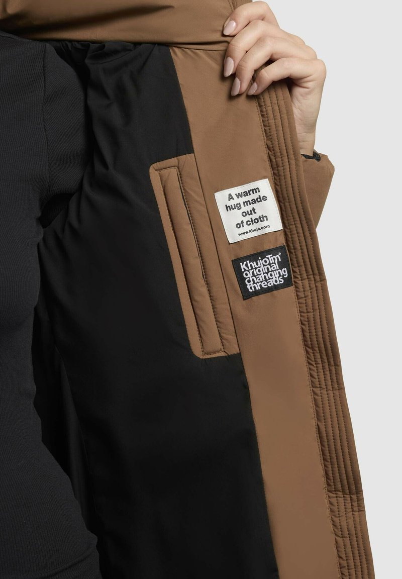 Braune gesteppte Jackeninnenseite mit schwarzem Futter, zeigt Stoffetiketten. Verfügt über einen geraden Schnitt und eine glatte Textur.