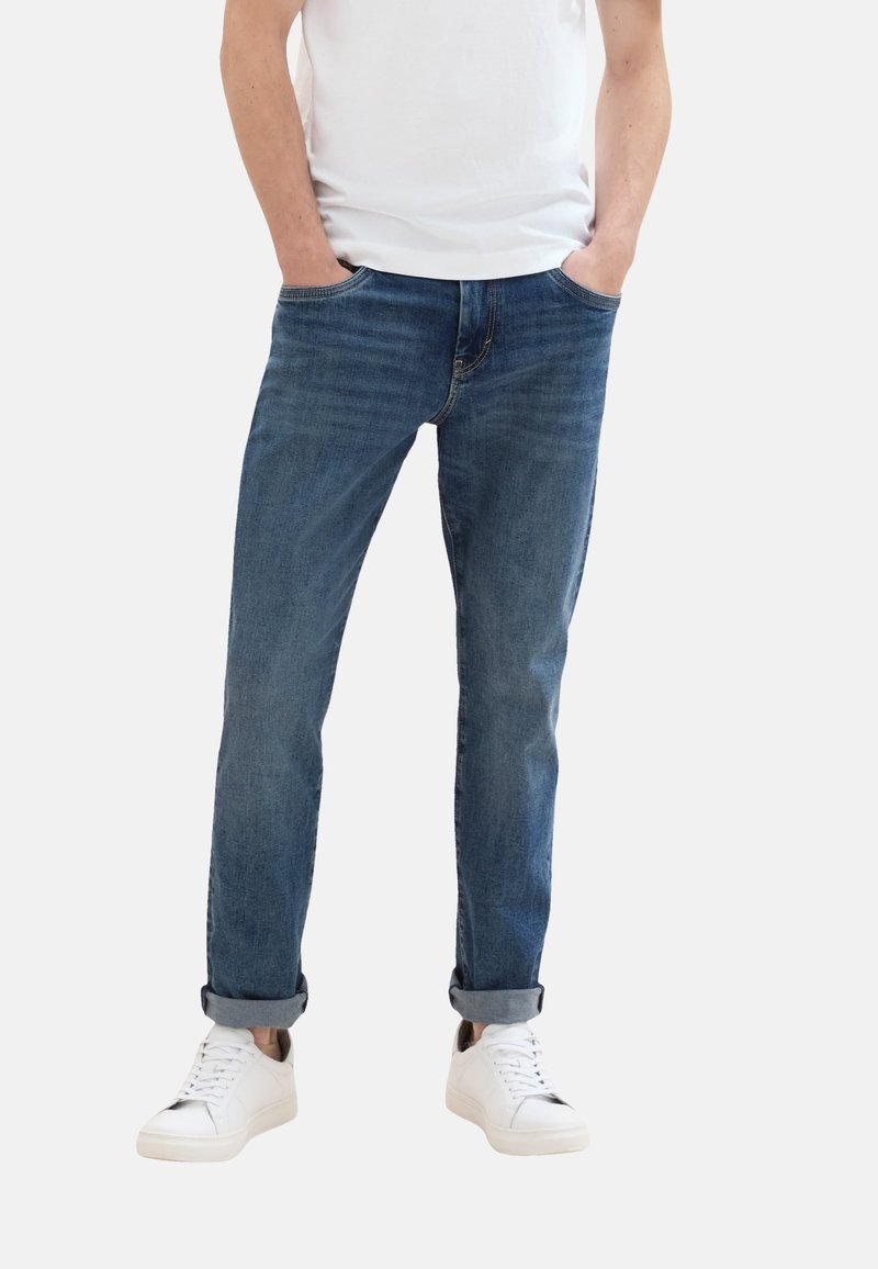 Jeans in denim blu con una vestibilità dritta, leggeri sbiadimenti e orli arrotolati, abbinati a sneakers bianche e una t-shirt bianca semplice.