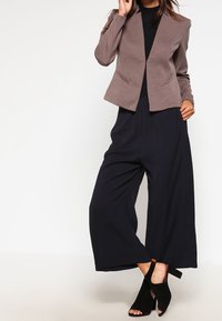 Femme portant un blazer marron texturé, un haut noir, un pantalon large noir et des talons noirs à bout ouvert, posant devant un fond blanc uni.