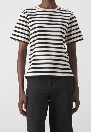 Person iført en kortærmet sort-og-hvidt vandretstribet skjorte med sorte bukser mod en ensfarvet baggrund.