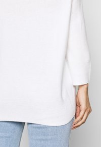 Main droite d'une personne portant un pull blanc côtelé et un jean bleu clair, avec la manche du pull se terminant au-dessus du poignet.