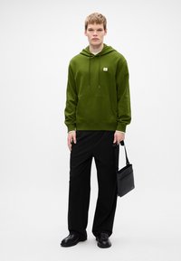 Hanorac verde măsliniu cu buzunar frontal și glugă cu șnur, asortat cu pantaloni negri largi și încălțăminte neagră, ținând o geantă de umăr neagră.