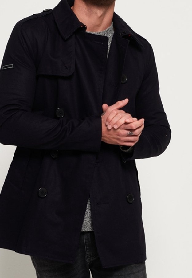 Mand iført en sort dobbeltradet frakke over en grå sweater, med hænderne foldet foran og en lille tatovering på venstre hånd.