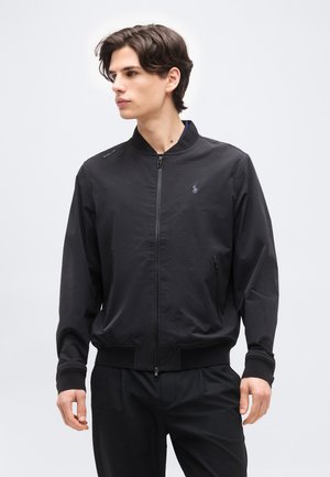 Jaunas vyras, dėvintis juodą striukę užsegamą užtrauktuku su mažu siuvinėtų logotipu kairėje krūtinės pusėje, stovintis priešais paprastą šviesią foną.