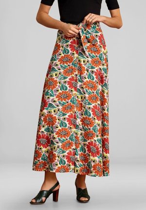 Kobieta wiążąca pasek w talii długiej kwiecistej spódnicy z pomarańczowymi, czerwonymi, żółtymi kwiatami i zielonymi liśćmi, ubrana w czarny top i ciemnozielone sandały na obcasie.