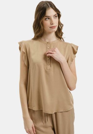 Młoda kobieta o falujących brązowych włosach, ubrana w bezrękawową beżową bluzkę na guziki i pasujące beżowe spodnie, patrząca lekko w prawo.