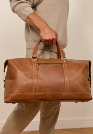 Hand die een grote bruine leren sporttas vasthoudt met ritsvak, beige trui en broek zichtbaar, effen achtergrond.