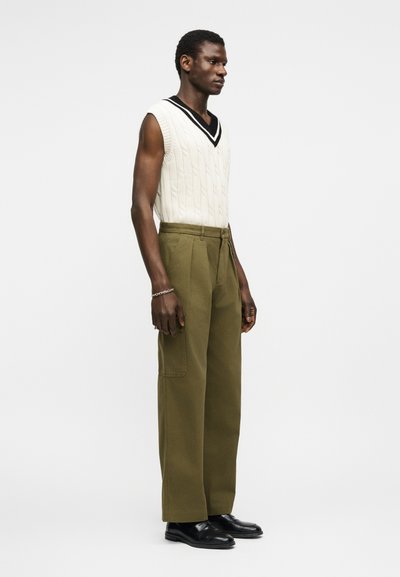 Άνδρας όρθιος που φοράει λευκό αμάνικο πουλόβερ με πλεκτό σχέδιο, παντελόνι φαρδύ σε ελιά πράσινο, μαύρα παπούτσια και ασημένιο βραχιόλι.