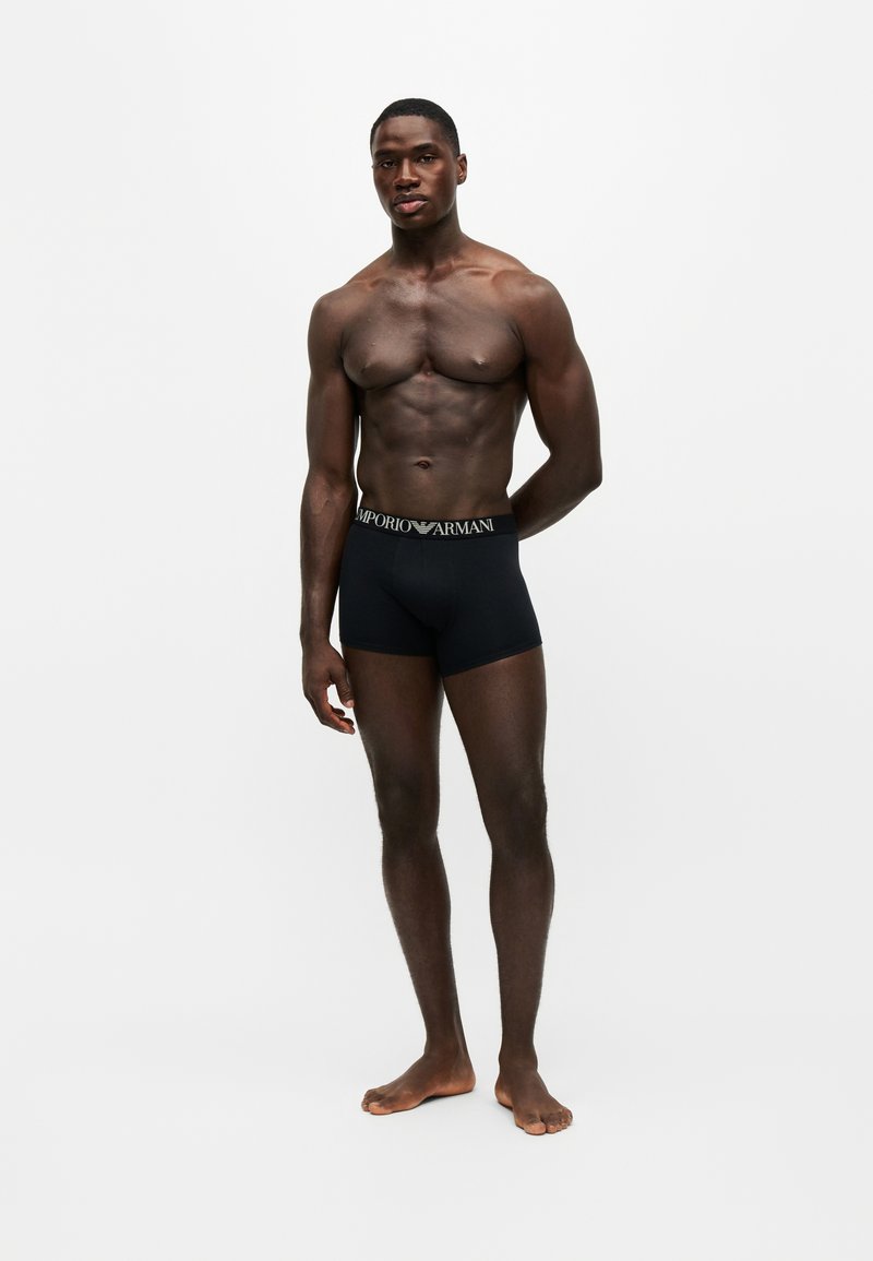 Homme musclé à la peau foncée, debout pieds nus, portant un boxer noir Emporio Armani sur un fond clair uni.