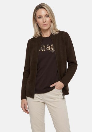 Donna con capelli biondi che indossa una giacca marrone scuro con zip, una maglietta nera stampata e pantaloni beige chiaro su uno sfondo uniforme.