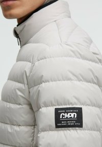 Hellegraue Pufferjacke mit gesteppten Abschnitten und einem hohen Kragen. Verfügt über ein schwarzes Etikett mit weißem Text am Oberarm. Glatte Textur.